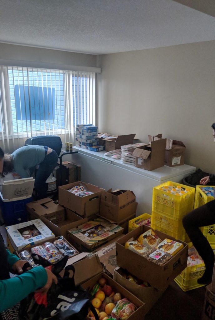 Several boxes on the floor of a white room full of food. Three people are sorting the food in the boxes.